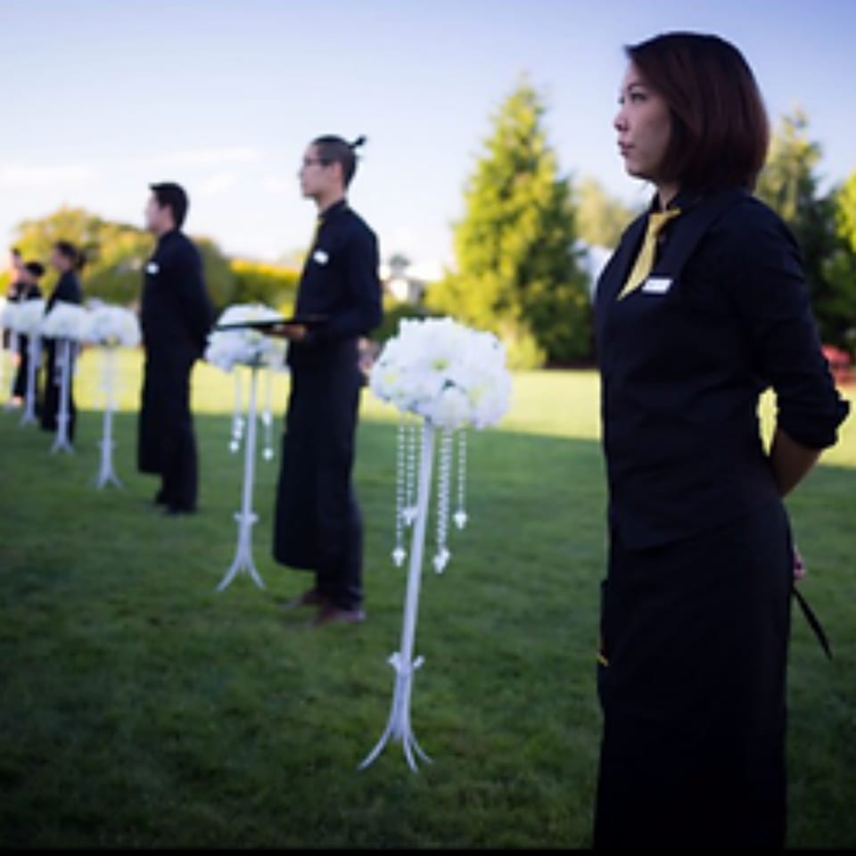 Caterers in black uniforms and yellow accents stand in a line on a grassy lawn with floral decorations.