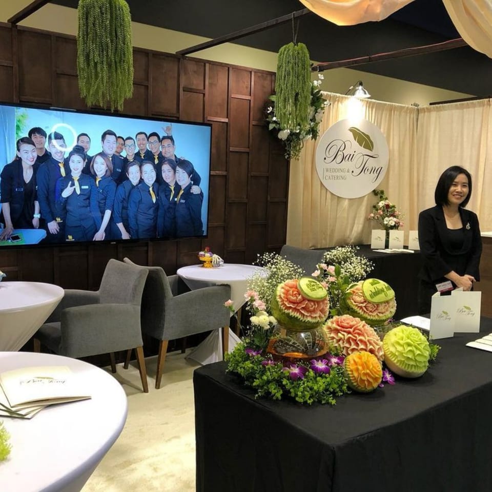 A Bai Tong Thai Street Cafe event booth with a woman, carved fruit, and a screen showing staff.