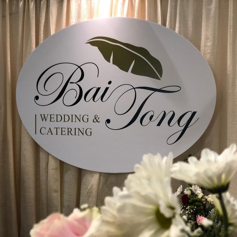 Bai Tong Wedding & Catering sign with a banana leaf logo, in front of beige curtains and flowers.