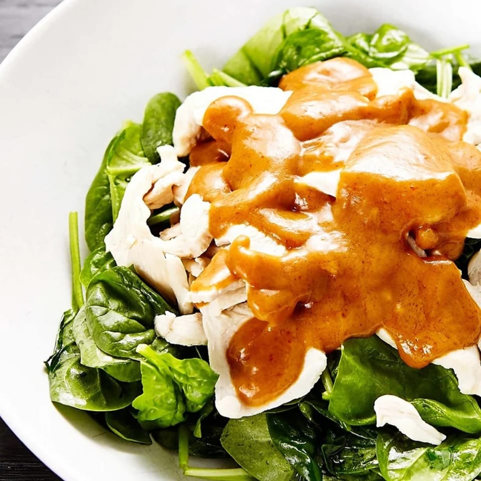 Spinach salad with sliced chicken and a rich peanut sauce in a white bowl.